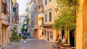 A narrow street with buildings on both sides, outdoor seating with tables and chairs, and potted plants along the walkway. - Appealing 2 Bedroom Villa, A/C, Private Pool, Panoramic Sea Views - In Nissaki, just 450m to Beach! (Nissaki)