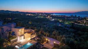Una casa moderna con piscina, rodeada de vegetación y con vistas a un pueblo costero al atardecer.