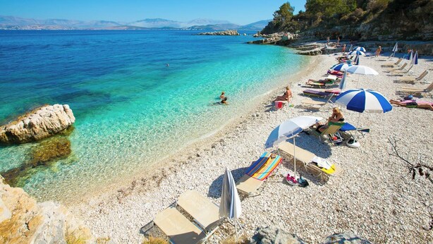 Una playa de guijarros con agua turquesa cristalina, afloramientos rocosos y una hilera de sillas y sombrillas de playa.