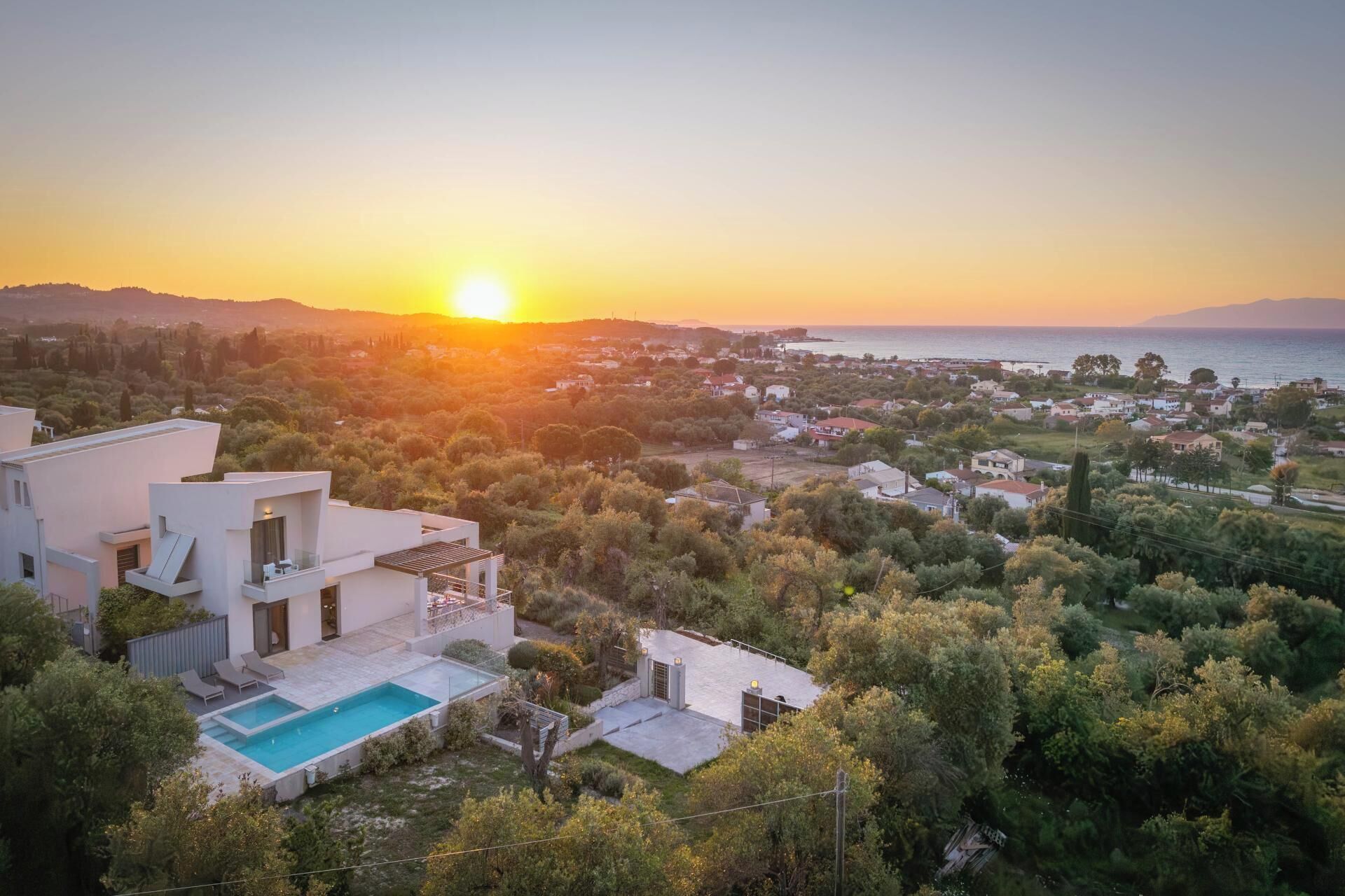 Une villa moderne dotée d’une piscine, entourée d’une végétation luxuriante et offrant une vue sur une ville côtière au coucher du soleil.