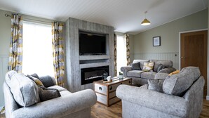 A living room with a fireplace, two grey sofas, a coffee table, and patterned curtains. - York Lodge Pet Friendly (East Sussex)
