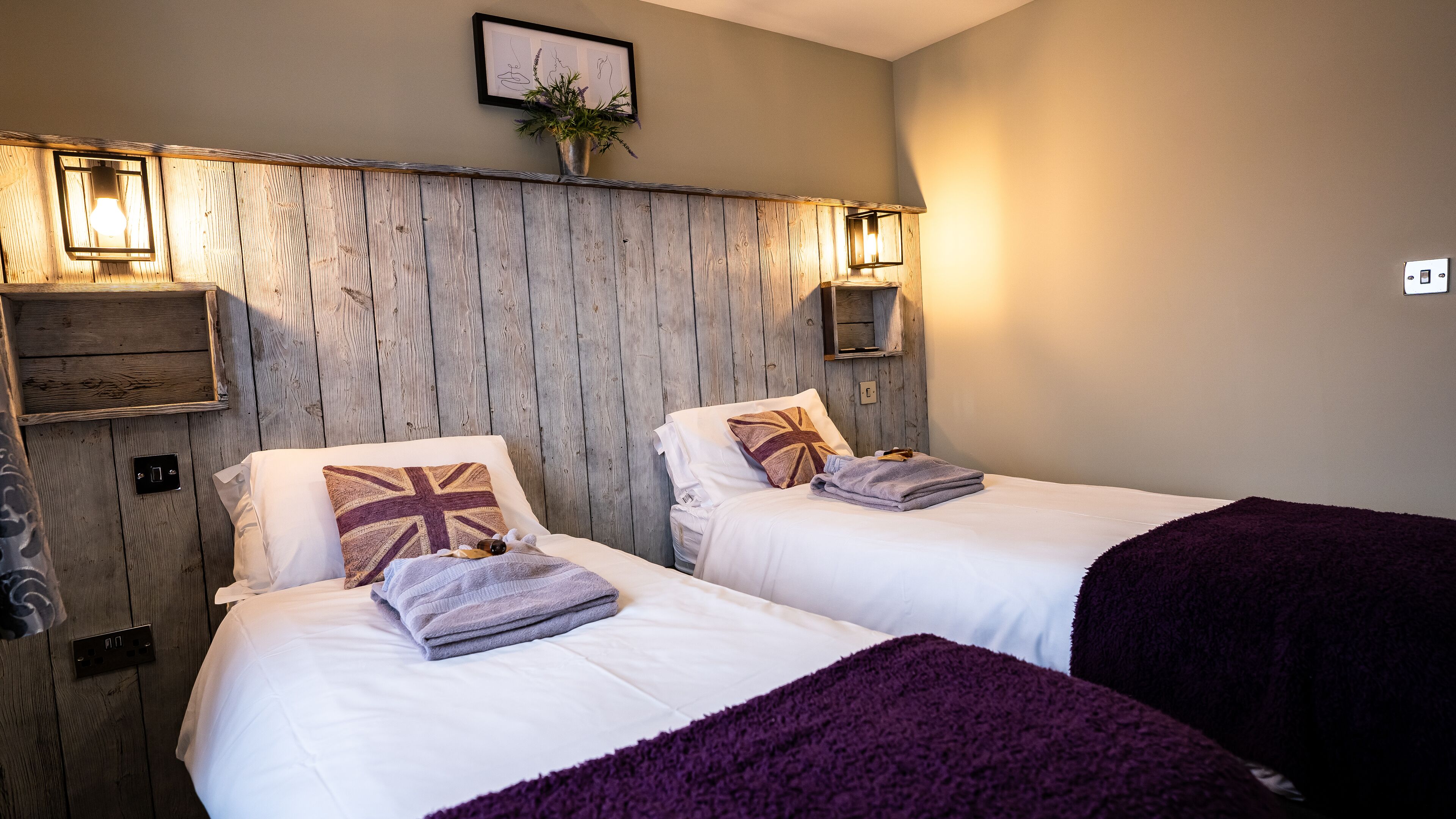 A room with two beds, wooden paneling on the wall, a framed picture, and a small shelf with a plant.