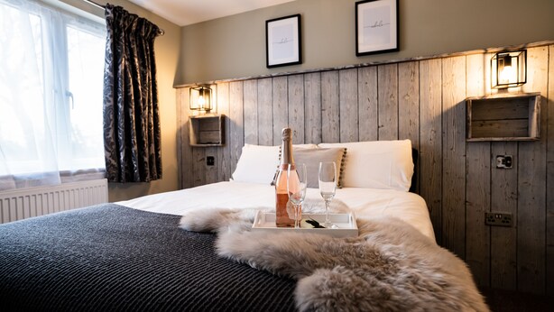 A hotel room with a wooden headboard, a bed with white linens, a fur throw, a tray with a bottle and glasses, and framed pictures on the wall. - York Lodge Pet Friendly (East Sussex)