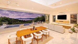 Una sala moderna con un televisor grande, una zona de comedor con mesa de madera y sillas blancas, y un balcón con vistas al paisaje.