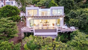 Una casa moderna de dos pisos con un balcón grande, rodeada de abundante vegetación.