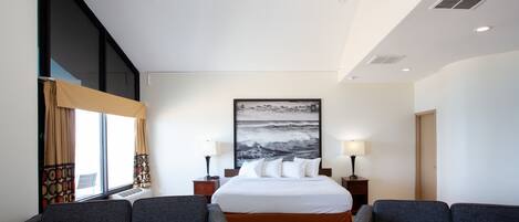 A hotel room with a large bed, two bedside tables with lamps, a chandelier, a large window with curtains, and a view of the ocean.