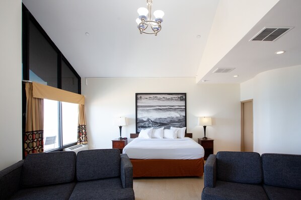 Quarto de hotel com uma cama grande, duas mesas de cabeceira com abajures, um candelabro, uma janela ampla com cortinas e vista para o oceano.