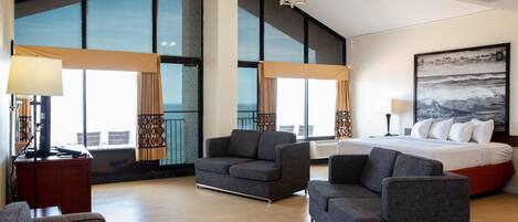 A modern hotel room with a large bed, two armchairs, a small table, and a large window offering a view of the city.
