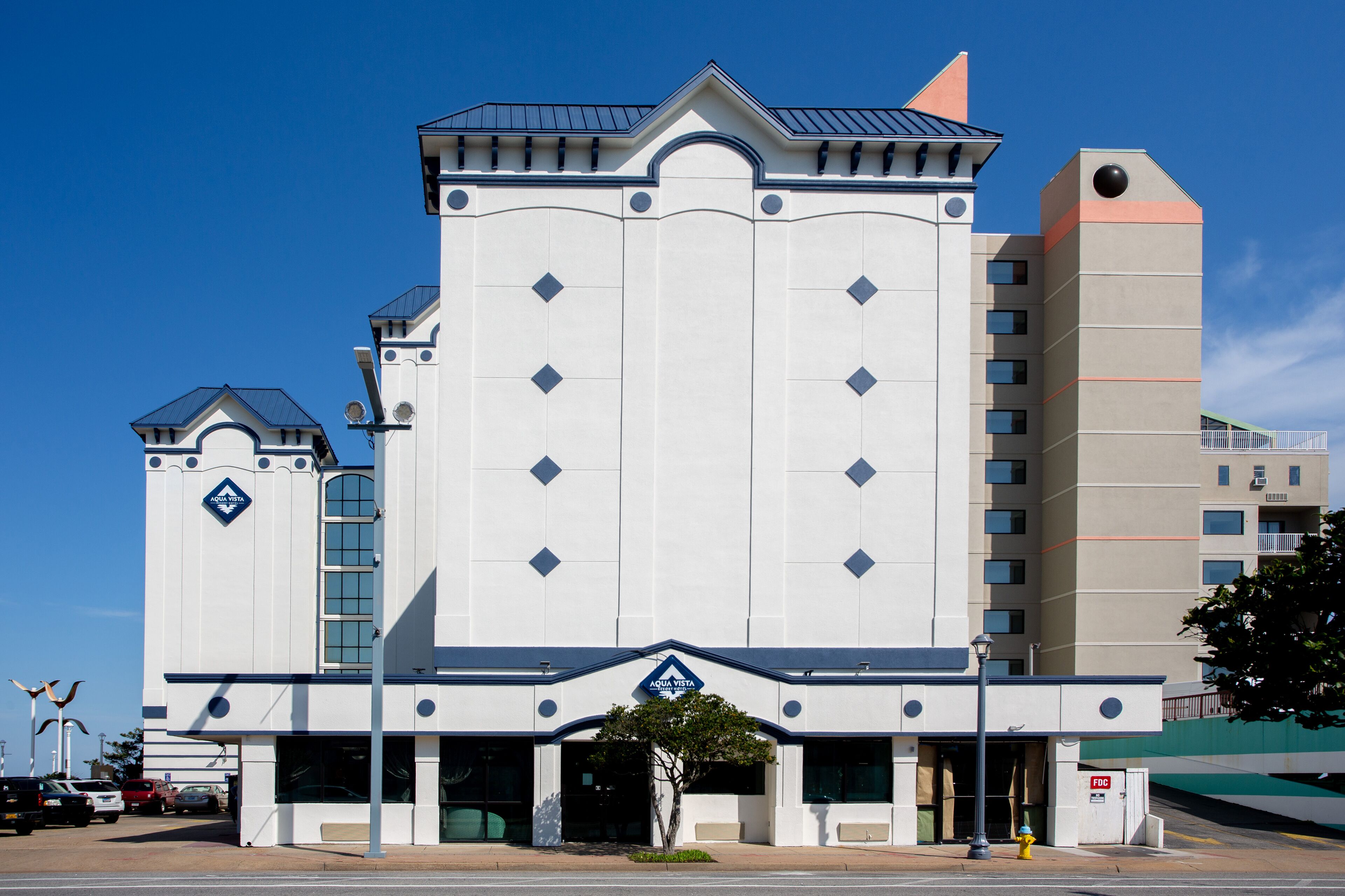 Un hotel de varios pisos con una entrada central, una estructura similar a una torre y un estacionamiento con varios automóviles.
