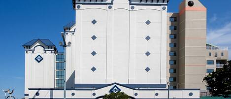 Un hotel de varios pisos con una entrada central, una estructura similar a una torre y un estacionamiento con varios automóviles.