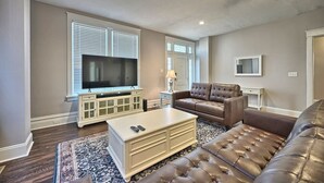 A living room with a large flat-screen TV, a brown leather sofa, a white coffee table, and a patterned rug. - Suites on Seneca - Lovely 2 Bed/1 Bath Apartment (Harrisburg)
