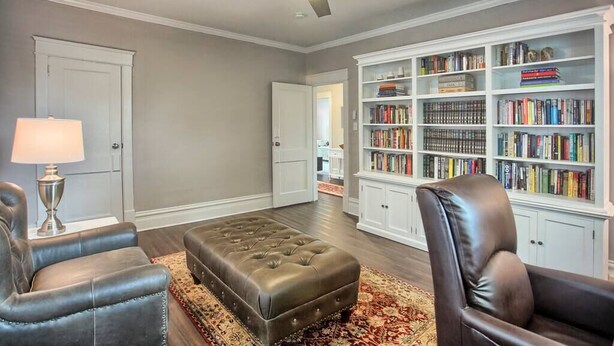 Una sala de estar con un otoman de cuero, dos sillones de cuero, un ventilador de techo y una estantería llena de libros.