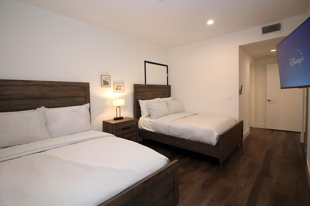 Une chambre d’hôtel avec deux lits, une table de chevet avec une lampe, un miroir et une porte ornée d’un logo Disney+.