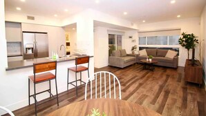 A modern kitchen with a built-in refrigerator, a dining area with a wooden table and chairs, a grey sofa, and a wooden floor.