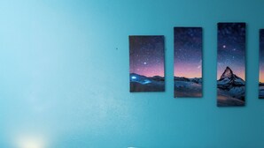 Un dormitorio con cama, almohadas blancas, mesita de noche con lámpara y cuatro cuadros en la pared.
