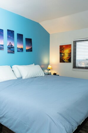 A bedroom with a large bed, white pillows, a bedside table with a lamp, and three wall art pieces depicting mountains and a sunset.