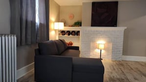 Un salón con un sofá de color oscuro, una chimenea de ladrillo blanco y un radiador.