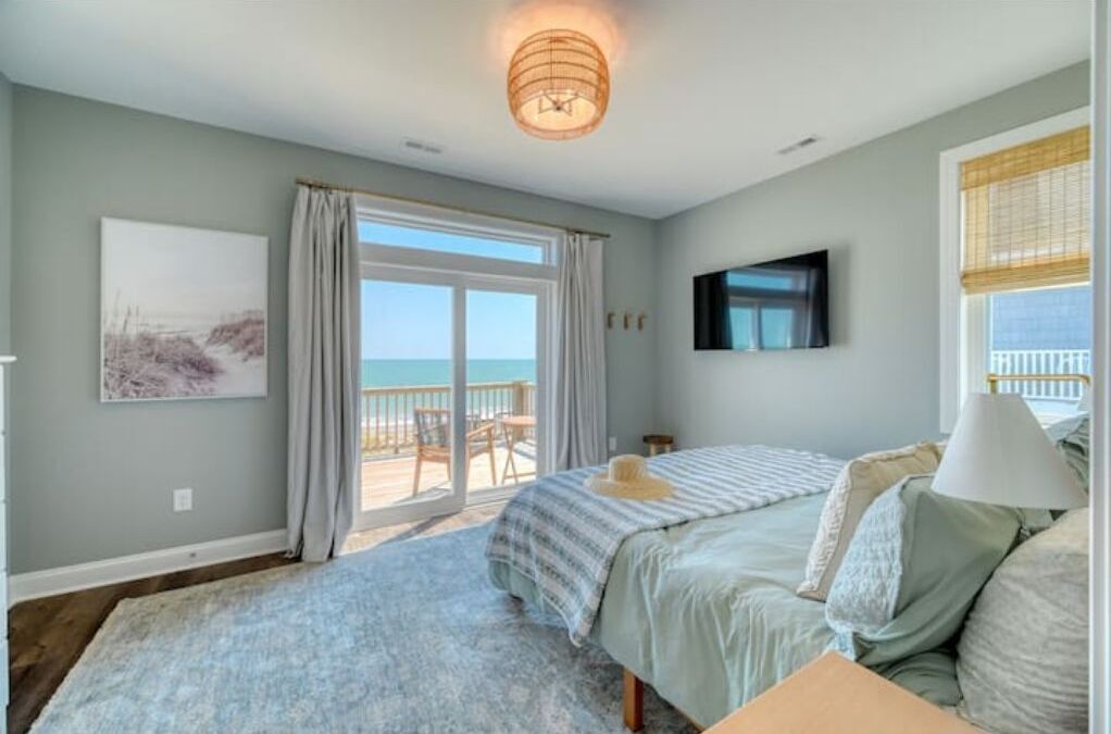 A bedroom with a bed, a TV, a window with curtains, and a view of the ocean.