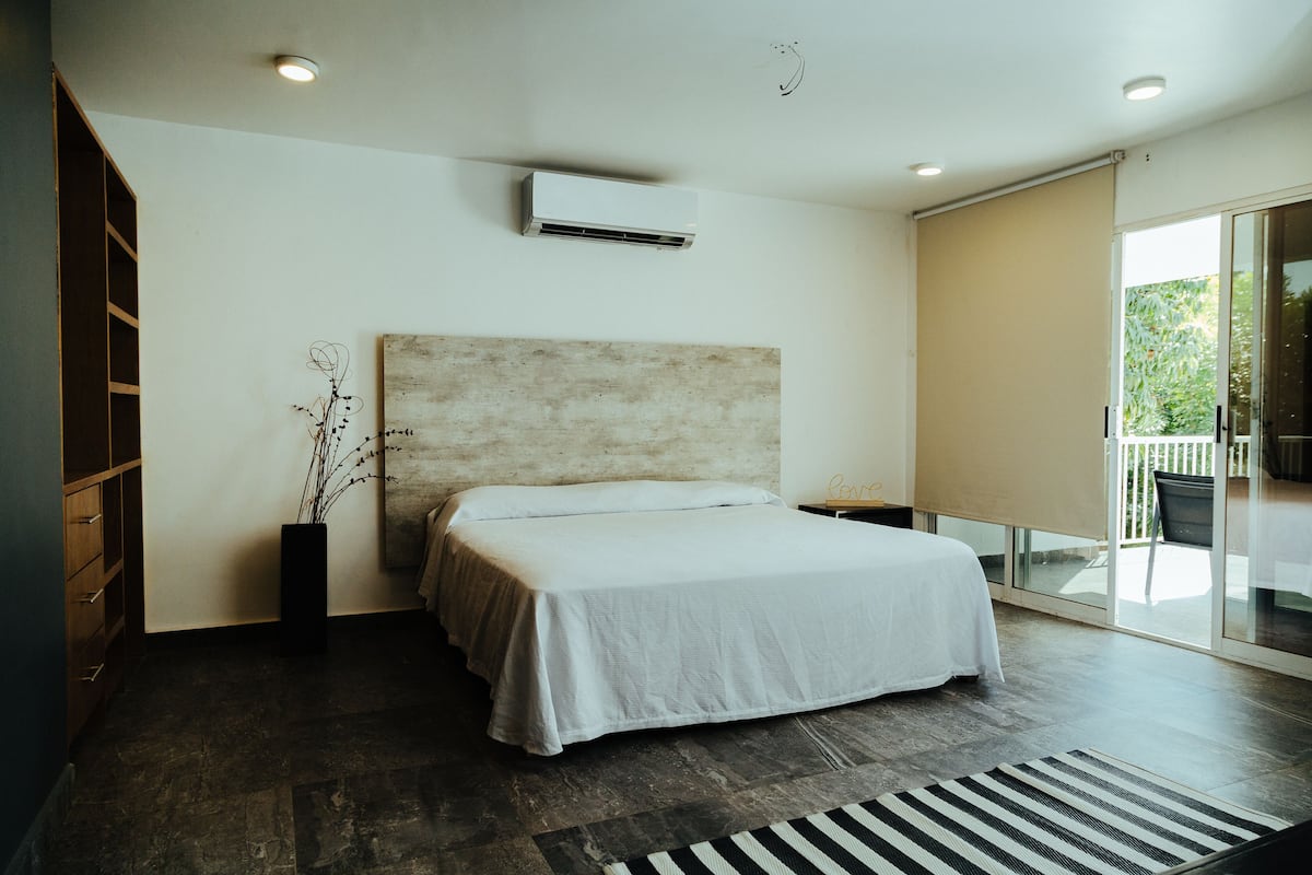 A modern bedroom with a large bed, a built-in wardrobe, a potted plant, and a sliding glass door to a balcony.