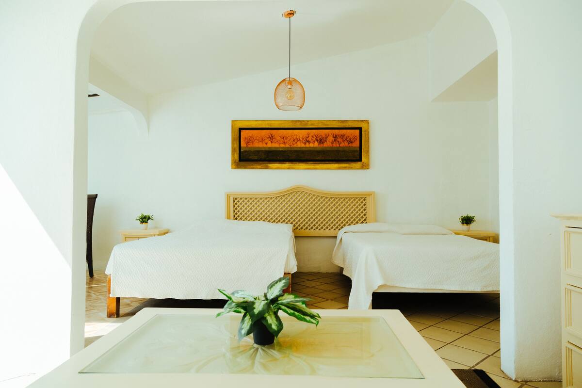 A hotel room with two beds, a painting on the wall, a hanging light fixture, and a small table with a plant.