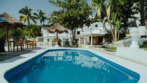 A swimming pool with a thatched umbrella, wooden chairs, and a stone wall. - Bendito Teques (Jojutla)