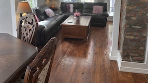 Un salon avec un canapé en cuir foncé, une table basse en bois et une table à manger avec des chaises. De grandes fenêtres aux cadres blancs et un mur en briques complètent la pièce.