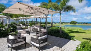 Eine Terrassenfläche mit Sitzgelegenheiten im Freien, darunter Korbstühle und ein großer Sonnenschirm, mit Blick auf einen Golfplatz und ein Gewässer.