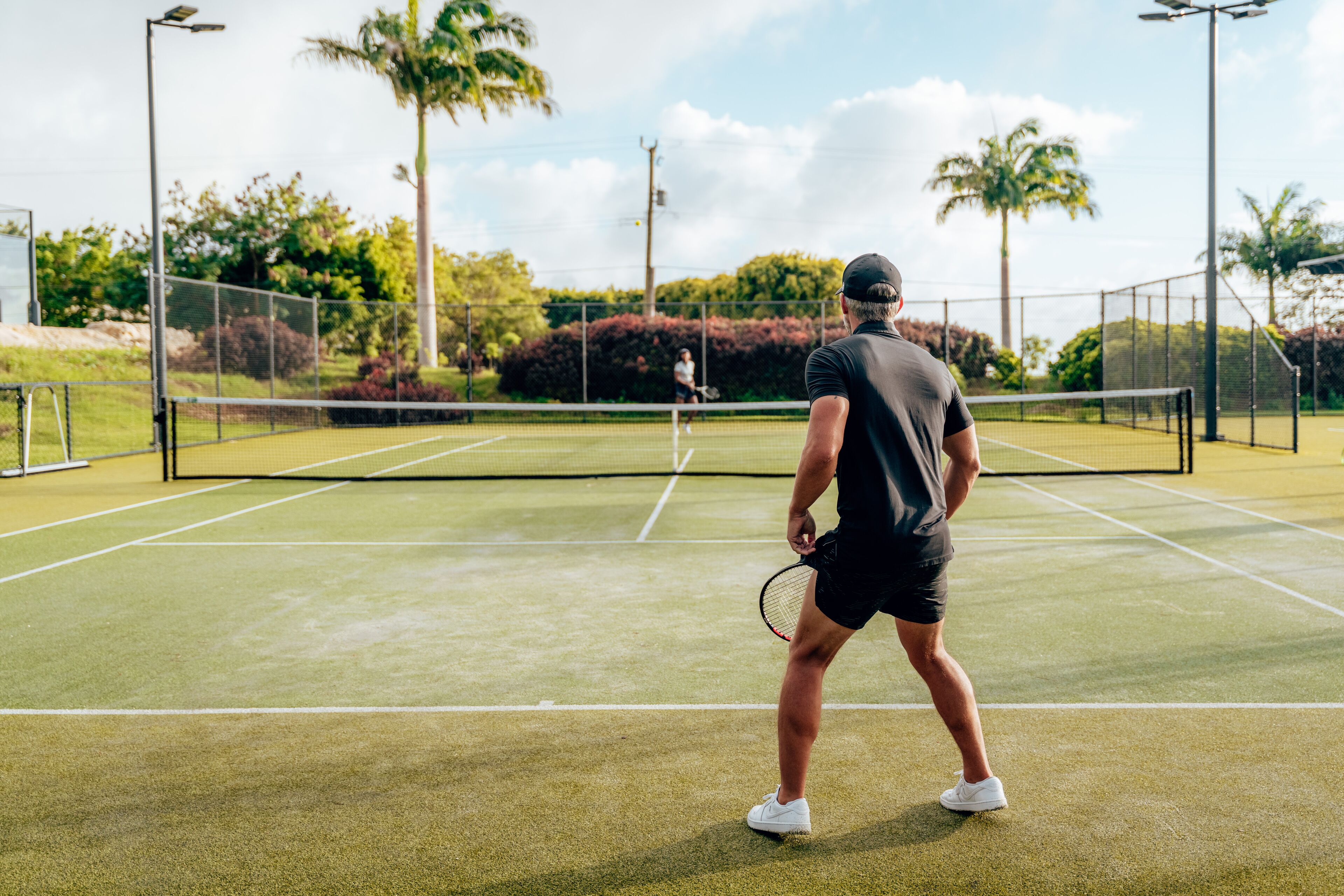A tennis court with a net, surrounded by greenery and palm trees.