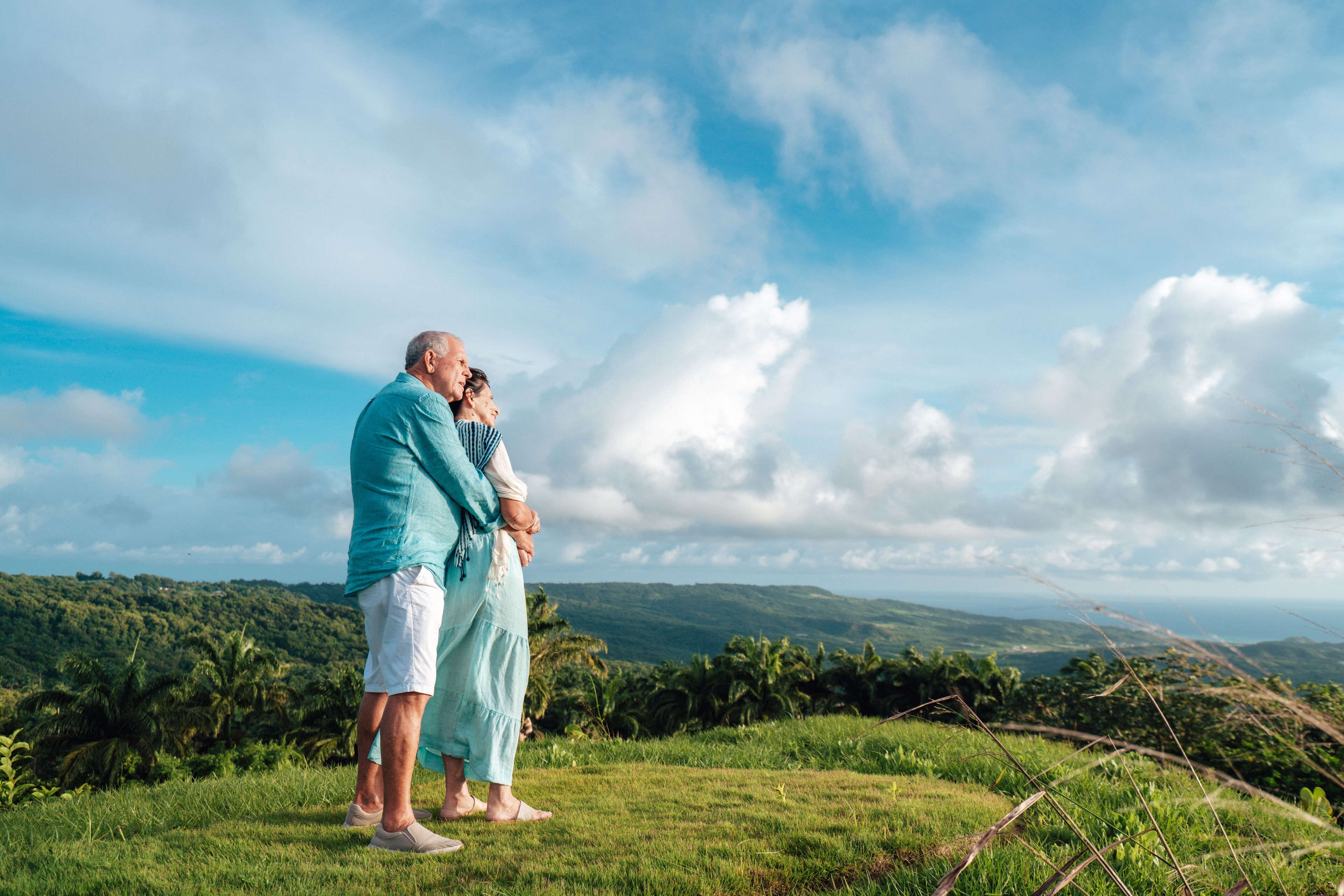 A couple standing on a grassy hill overlooking a lush landscape with hills and a clear sky.