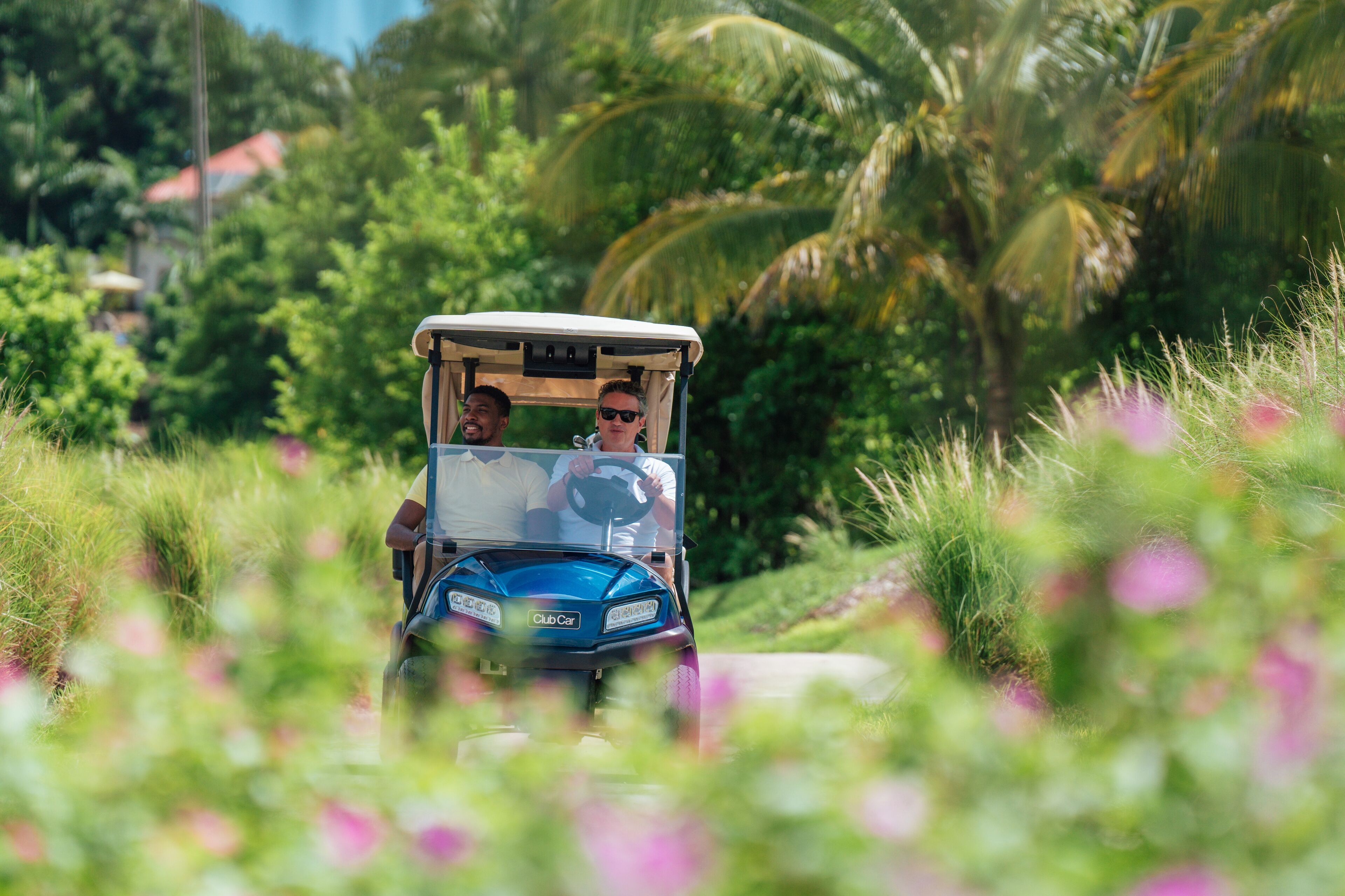 A golf cart driving through a lush, green area with palm trees and flowers in the foreground.