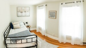 A bedroom with a bed, a nightstand, a lamp, and two windows with sheer curtains. - Serenity in South Wedge: desirable walking community meets vibrant tranquillity! (Rochester)