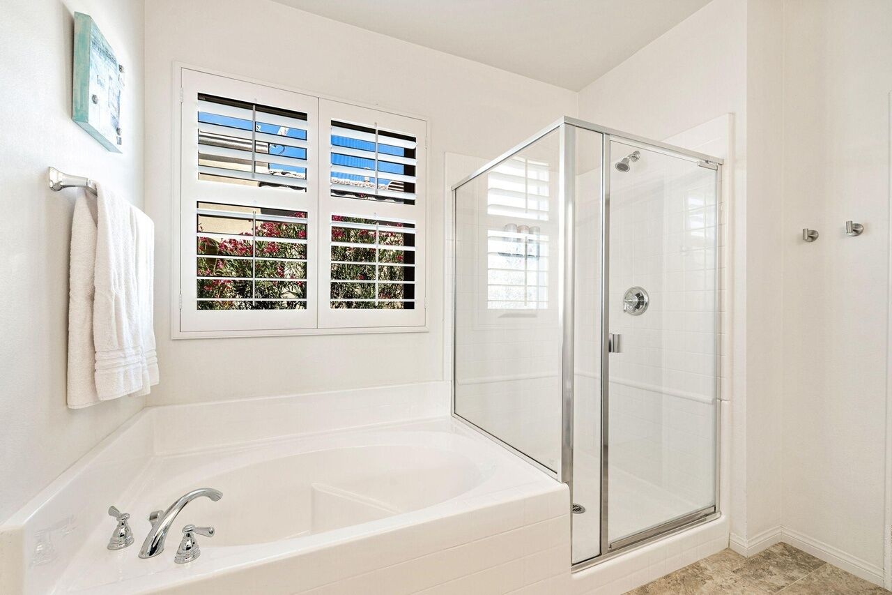 Un baño moderno con ducha envuelta en vidrio, una bañera independiente y una ventana con persianas decorativas.