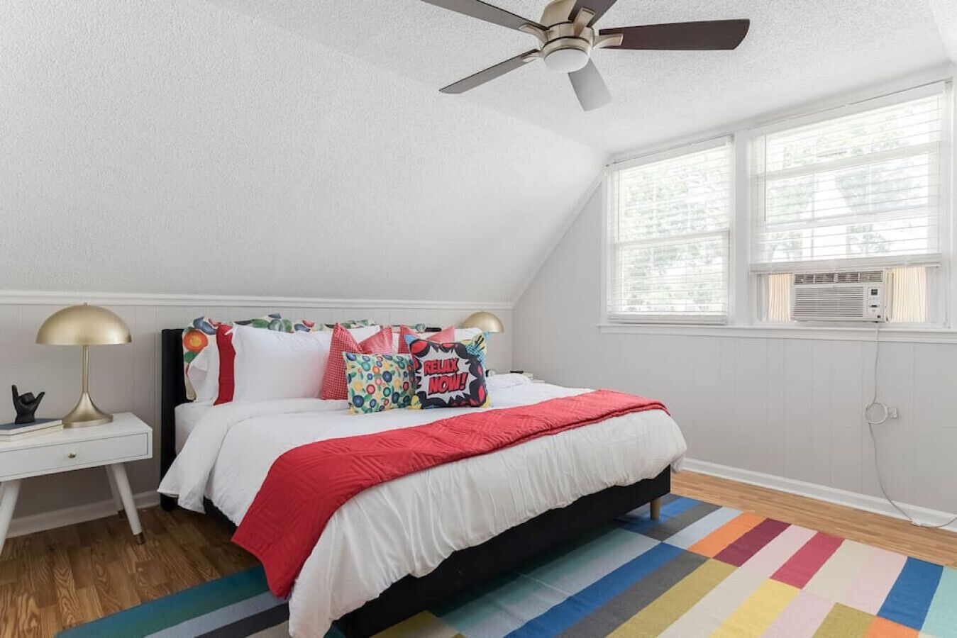 A bedroom with a bed, a nightstand, a ceiling fan, a window with blinds, and a colorful striped rug.