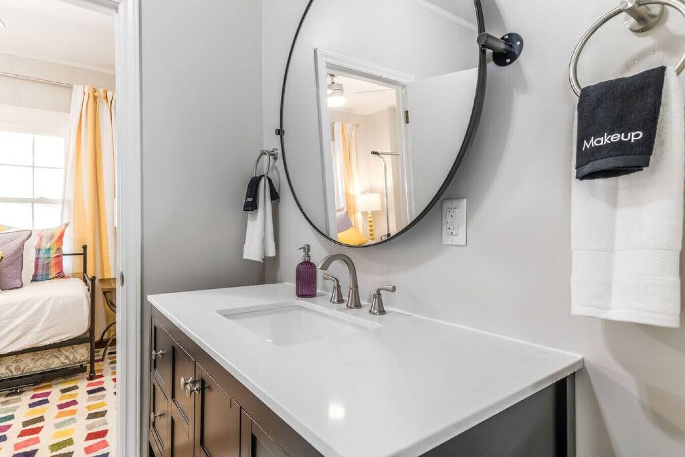 Une salle de bain moderne avec un lavabo blanc, un miroir rond et un porte-serviettes sur lequel est posée une serviette noire étiquetée « Maquillage ».