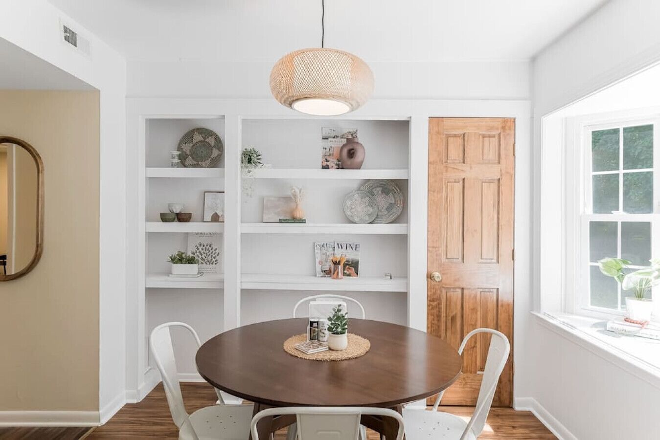 A dining area with a round table, white chairs, a wooden door, and a window with a view.