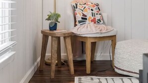 A corner room with a floor lamp, a small table with a plant, a colorful pillow, and a white bed.