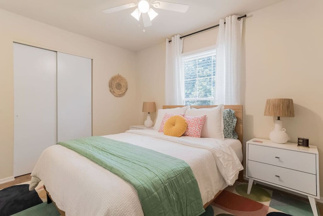 Un dormitorio con cama, mesitas de noche, una ventana con cortinas, un ventilador de techo y un adorno en la pared.