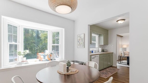 A dining area with a round table and chairs, a window with blinds, and a view of greenery outside.