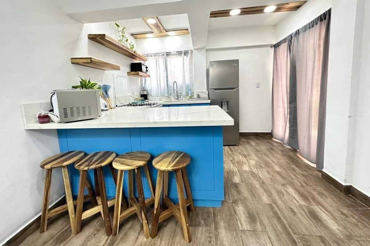 A modern kitchen with a white countertop, blue accent, wooden stools, and built-in appliances.