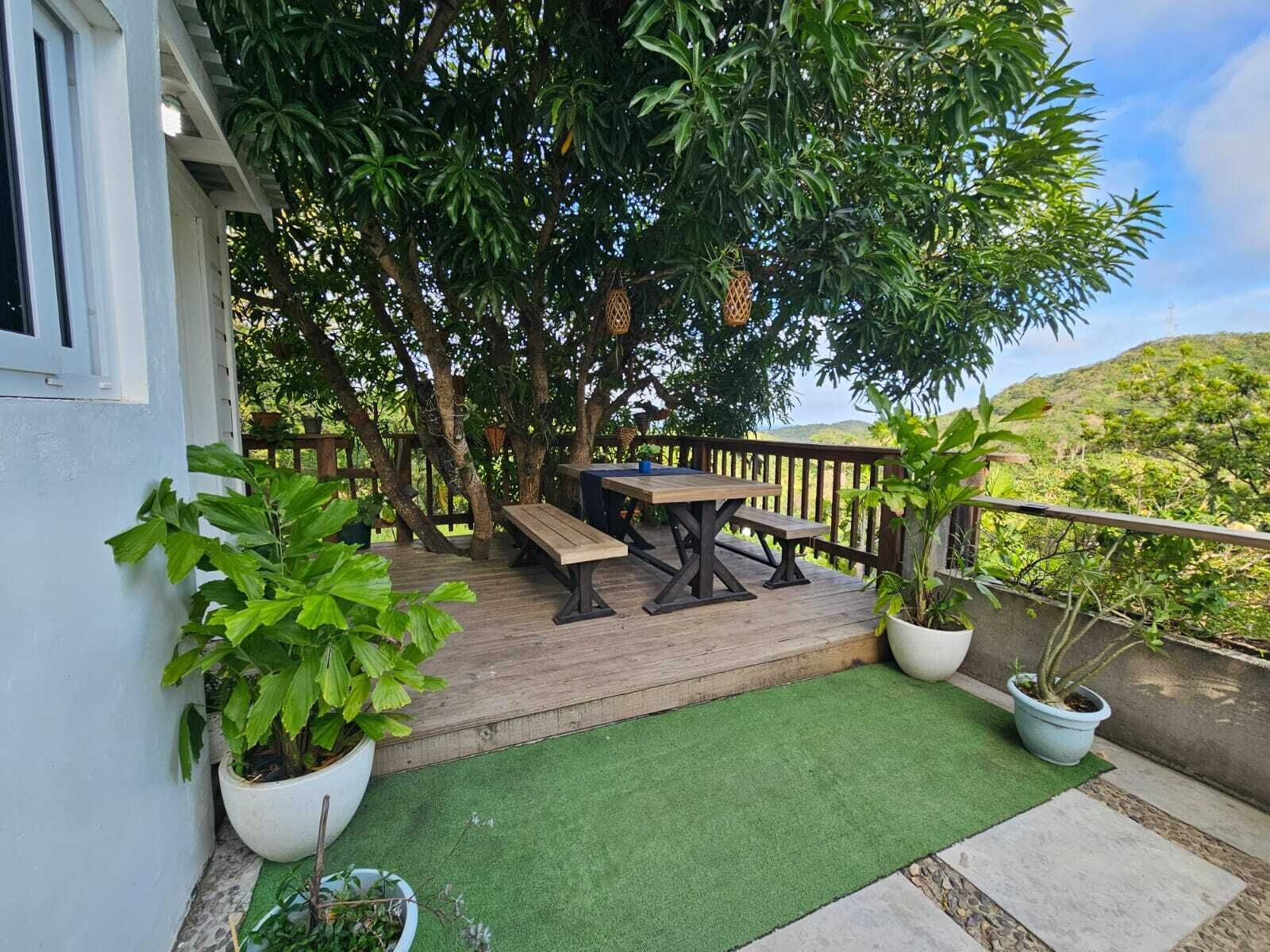 A wooden deck with a table and benches, surrounded by potted plants and a view of a hilly landscape.