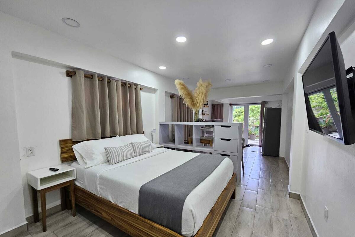 A modern bedroom with a wooden bed, a bedside table, a built-in TV, and a view of the outdoors.