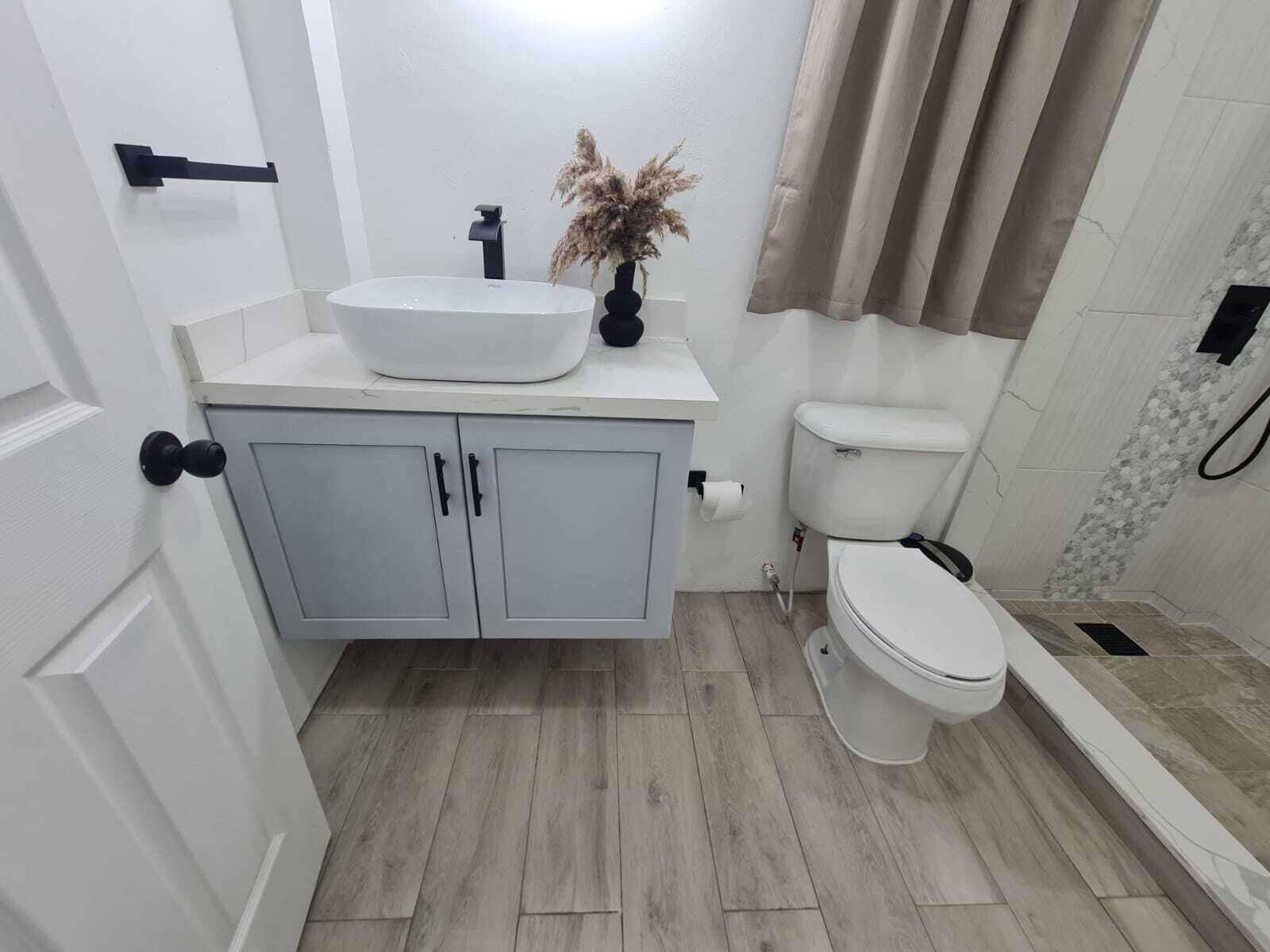 A bathroom with a white sink, grey cabinets, a toilet, and a shower area.