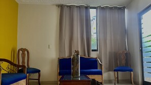 Uma sala de estar com um sofá azul, uma cadeira de madeira, uma mesa pequena de madeira e uma janela grande com cortinas.