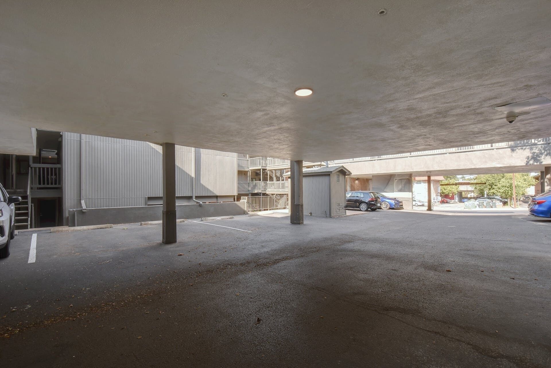 Un estacionamiento vacío debajo de un edificio con techo plano, pilares de concreto y un pequeño cobertizo.