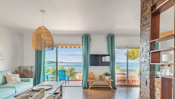 Un vaste salon doté d’une grande fenêtre offrant une vue sur la mer, un coin salon confortable avec un canapé et des fauteuils, ainsi qu’une table basse en bois.