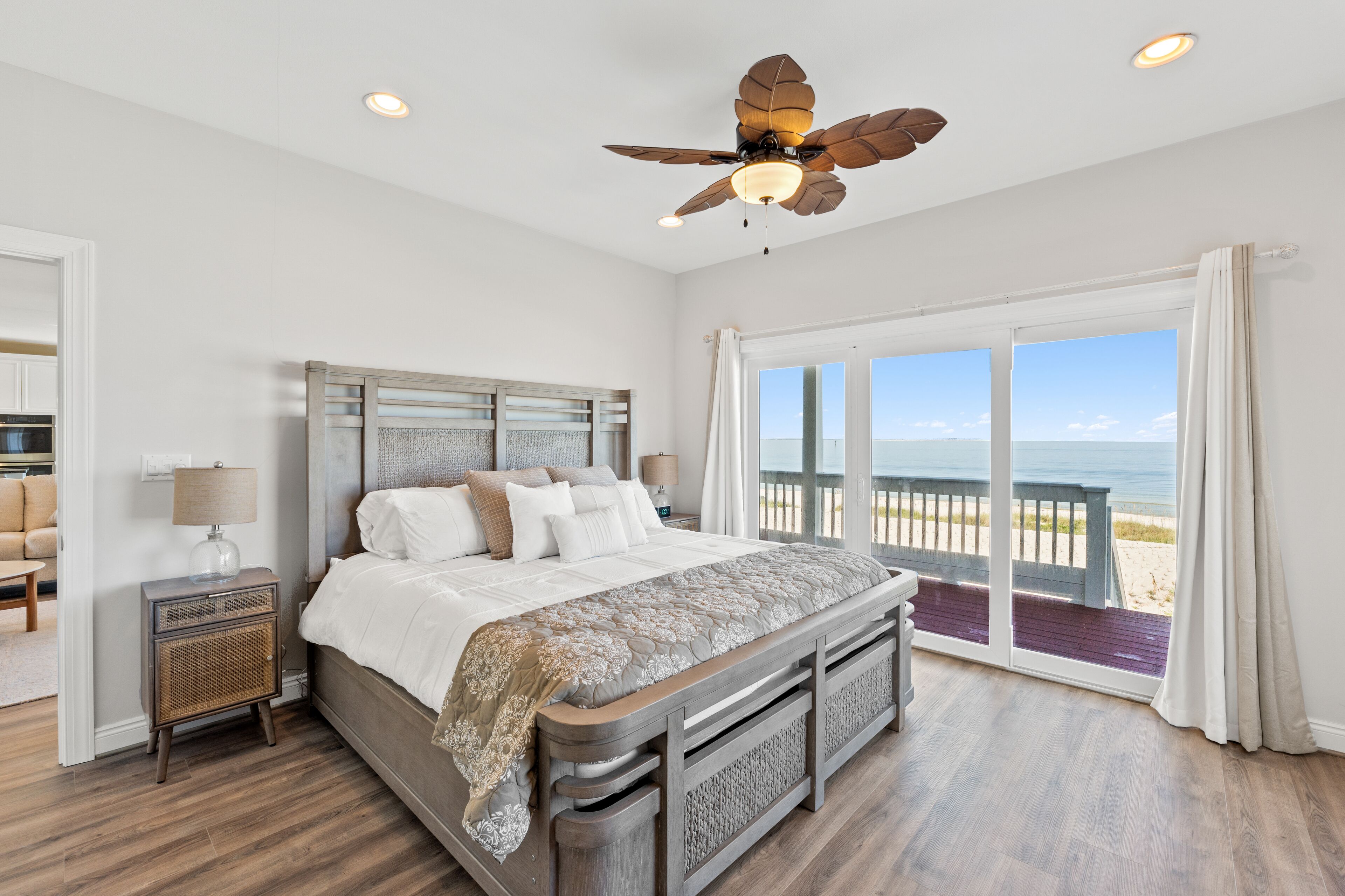 A bedroom with a large bed, a nightstand, a ceiling fan, and a view of the beach through large windows.