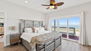Una camera da letto con un letto grande, un comodino, una ventola a soffitto e vista sulla spiaggia attraverso ampie finestre.