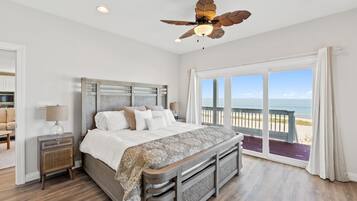A bedroom with a large bed, a nightstand, a ceiling fan, and a view of the beach through large windows.