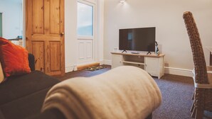 Ein Wohnzimmer mit einer Holztür, einem Fernseher auf einem weißen Schrank, einem braunen Sessel und einem gewebten Stuhl.
