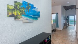 Een gang met een witte, gestructureerde wand, een zwarte console tafel en drie ingelijste strandschilderijen.